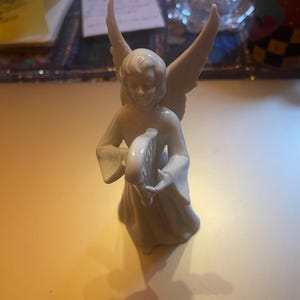 May include: A cream-colored angel figurine with outstretched wings, holding a crescent moon-shaped object. The angel is wearing a flowing robe. The figurine is on a yellow surface.