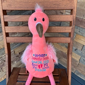 May include: A pink plush flamingo toy with a brown beak and black eyes. The flamingo has embroidered details including a name, birthdate, weight (3.6 kg), and height (48 cm). It is sitting on a wooden chair.