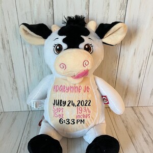 May include: A white plush cow with black patches, ears, and hooves. The cow has embroidered details, including a pink tongue, and text that reads "Adalynne Jo Harmon, July 24, 2022, 5 lbs, 15 oz, 19 3/4 inches, 6:33 PM."