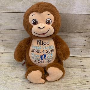 May include: A brown plush monkey with embroidered details. The monkey has a tan face, large eyes, and a smiling expression. The monkey's chest features embroidered text with a name, date, and measurements. The monkey is approximately 23 inches tall.