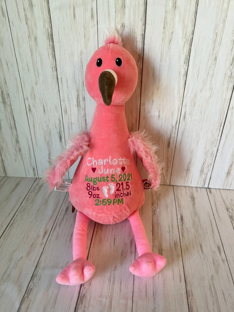 May include: A pink plush flamingo with a brown beak and black eyes. The flamingo has embroidered text that reads "Charlotte June August 5, 2021 8lbs 9oz 54.6 cm 2:59 PM". The flamingo has fluffy wings and legs.
