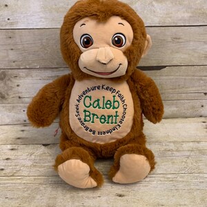 May include: A plush brown monkey with a tan face, ears, and hands. The monkey has embroidered eyes and a smiling mouth. The chest features the name "Caleb Brent" in green embroidery, surrounded by the words "Seek Adventure, Keep Faith, Choose Kindness, Be Brave."
