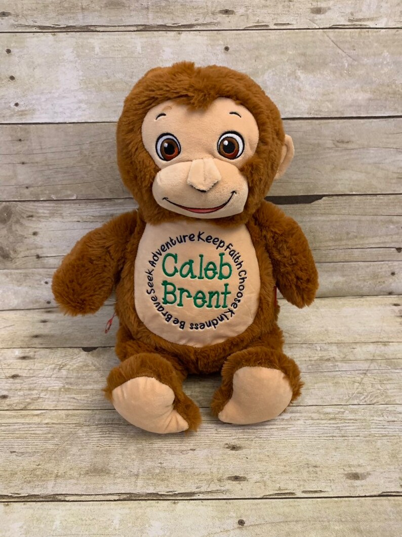 May include: A plush brown monkey with a tan face, ears, and hands. The monkey has embroidered eyes and a smiling mouth. The chest features the name "Caleb Brent" in green embroidery, surrounded by the words "Seek Adventure, Keep Faith, Choose Kindness, Be Brave."