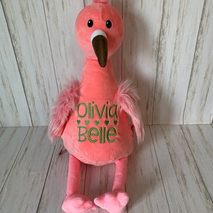 May include: A soft pink flamingo toy with a brown beak, black eyes, and green embroidered text reading "Olivia Belle" with green heart accents. It has fluffy pink wing feathers and a pink head crest. The toy is made of plush material.