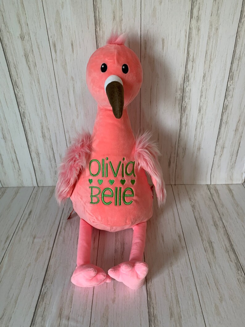 May include: A soft pink flamingo toy with a brown beak, black eyes, and green embroidered text reading "Olivia Belle" with green heart accents. It has fluffy pink wing feathers and a pink head crest. The toy is made of plush material.