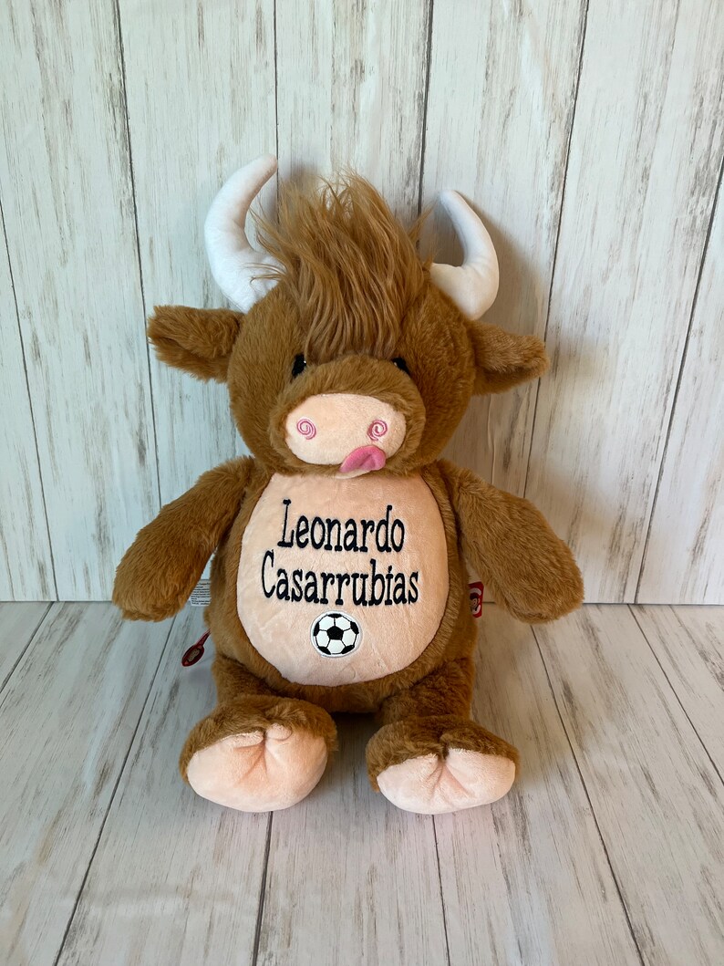 May include: A brown plush bull with white horns and a tuft of brown fur. The bull has a pink tongue sticking out and the name "Leonardo Casarrubias" embroidered on its belly, along with a soccer ball.