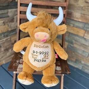May include: A plush, brown Highland cow stuffed animal with white horns and a pink tongue sticking out. The cow has embroidered details, including a name, birthdate, weight (9 lbs 5 oz), length (23 inches), and time of birth.