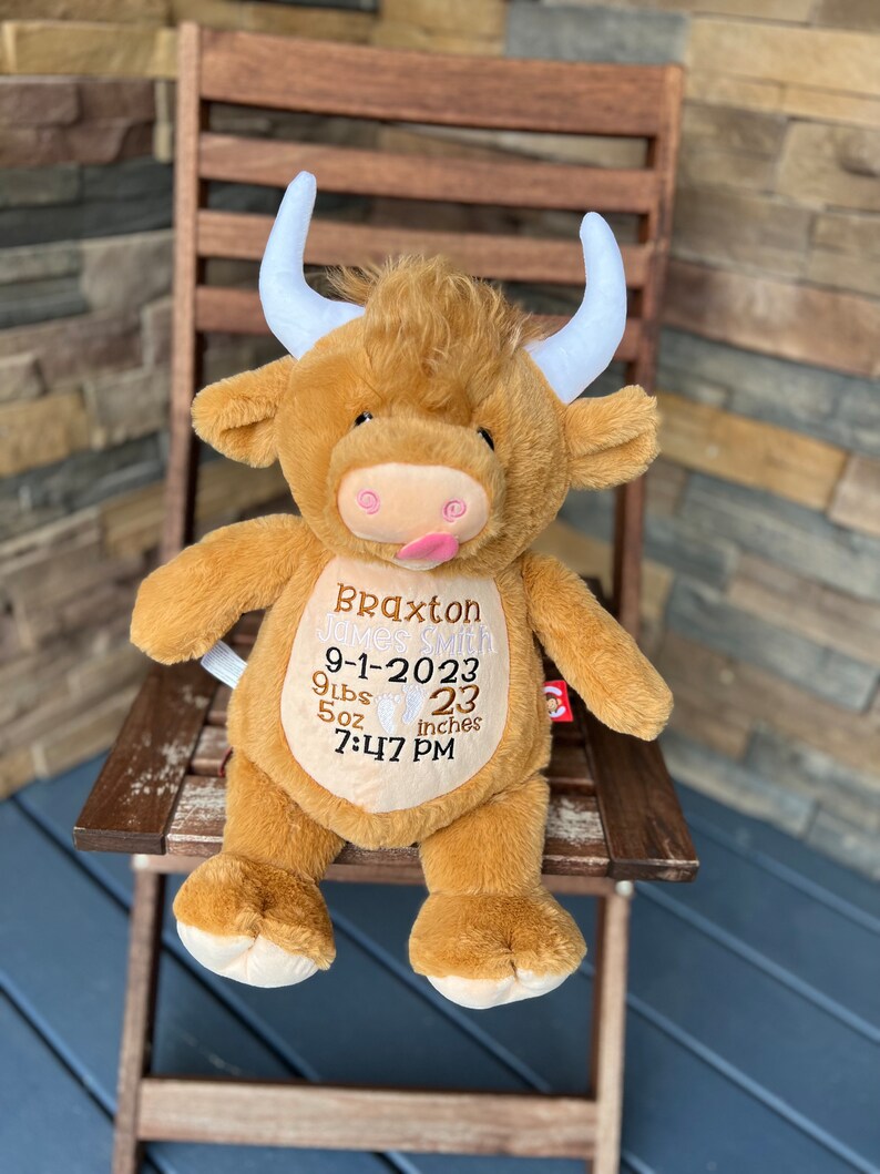 May include: A plush, brown Highland cow stuffed animal with white horns and a pink tongue sticking out. The cow has embroidered details, including a name, birthdate, weight (9 lbs 5 oz), length (23 inches), and time of birth.