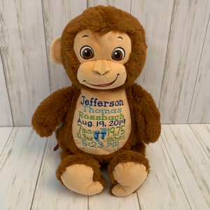May include: A plush brown monkey with embroidered details. The monkey has a tan face, ears, and feet. The monkey's chest features embroidered text in blue and green, including a name, date, weight, and length. The monkey is sitting upright.