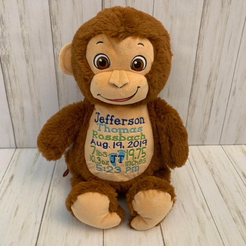 May include: A plush brown monkey with embroidered details. The monkey has a tan face, ears, and feet. The monkey's chest features embroidered text in blue and green, including a name, date, weight, and length. The monkey is sitting upright.