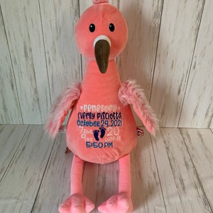 May include: A pink plush flamingo with a brown beak and black eyes. The flamingo has embroidered text in blue, including a name, date, time, weight (3.4 kg), and length (51 cm). The flamingo has fluffy wings and legs.
