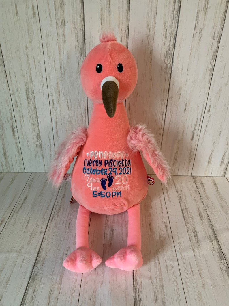 May include: A pink plush flamingo with a brown beak and black eyes. The flamingo has embroidered text in blue, including a name, date, time, weight (3.4 kg), and length (51 cm). The flamingo has fluffy wings and legs.