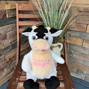 May include: A plush cow toy with black and white markings, sitting on a wooden chair. The cow has a pink tongue and the name "Teagan" embroidered in pink on its chest. The background includes a brick wall and greenery.