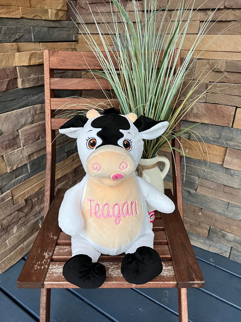 May include: A plush cow toy with black and white markings, sitting on a wooden chair. The cow has a pink tongue and the name "Teagan" embroidered in pink on its chest. The background includes a brick wall and greenery.