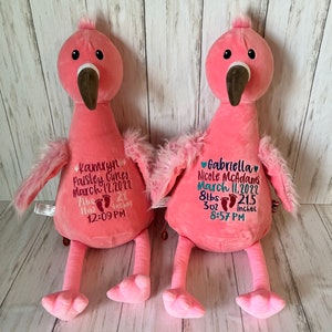 May include: Two pink plush flamingo toys with embroidered details. Each flamingo has a brown beak, black eyes, and fluffy wings. One flamingo has the name "Kamryn Paisley Cuneo" and birth details embroidered on its chest. The other has "Gabriella Nicole McAdams" and birth details.