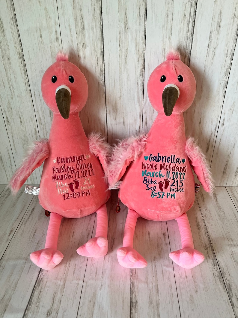 May include: Two pink plush flamingo toys with embroidered details. Each flamingo has a brown beak, black eyes, and fluffy wings. One flamingo has the name "Kamryn Paisley Cuneo" and birth details embroidered on its chest. The other has "Gabriella Nicole McAdams" and birth details.