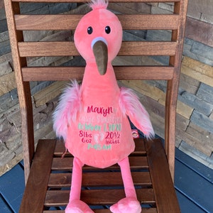 May include: A pink plush flamingo toy with a brown beak and black eyes. The flamingo has pink feathers and embroidered text that includes a name, date, weight (3.8 kg), and height (52 cm). The toy is sitting on a wooden chair.