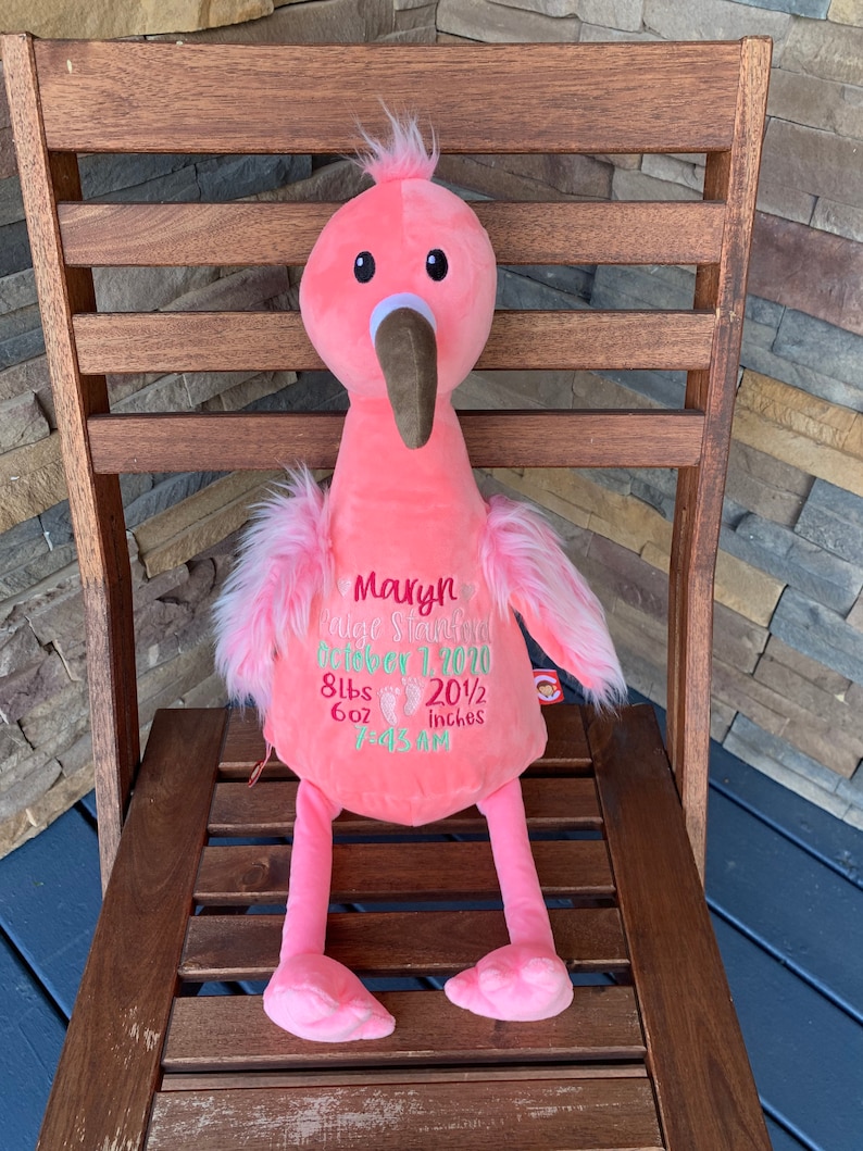 May include: A pink plush flamingo toy with a brown beak and black eyes. The flamingo has pink feathers and embroidered text that includes a name, date, weight (3.8 kg), and height (52 cm). The toy is sitting on a wooden chair.