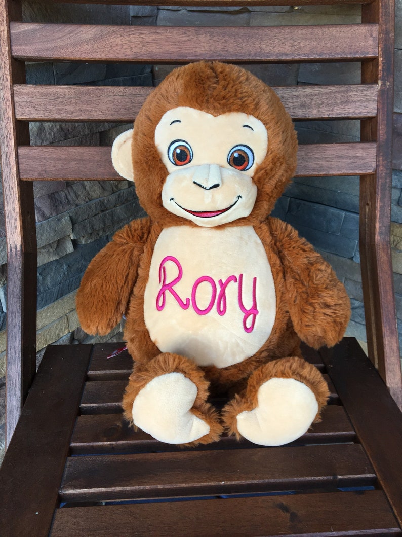 May include: A plush brown monkey with a cream-colored face, hands, and feet. The monkey has embroidered eyes and the name "Rory" in pink script on its chest. It is sitting on a wooden chair.