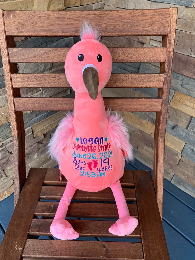 May include: A pink plush flamingo toy with a brown beak and black eyes. The flamingo has embroidered details including a name, birthdate, weight (3.6 kg), and height (48 cm). It is sitting on a wooden chair.