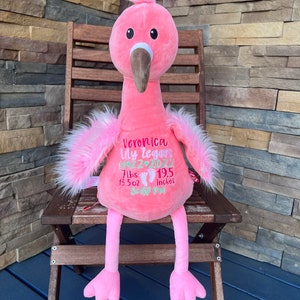 May include: A pink plush flamingo toy with a long neck, brown beak, and fluffy pink feathers. The flamingo has embroidered details, including a name, date, weight (3.18 kg, 383 g), and height (49.5 cm). It is sitting on a wooden chair.