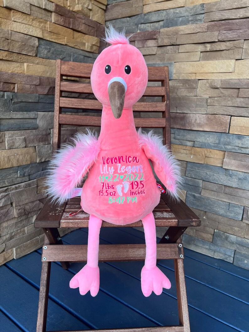 May include: A pink plush flamingo toy with a long neck, brown beak, and fluffy pink feathers. The flamingo has embroidered details, including a name, date, weight (3.18 kg, 383 g), and height (49.5 cm). It is sitting on a wooden chair.
