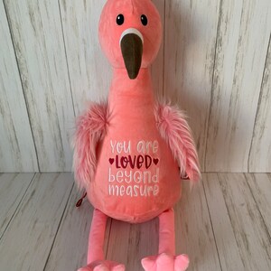 May include: A soft pink flamingo toy with a long neck, brown beak, and fluffy pink feathers. The flamingo has the embroidered text "You are loved beyond measure" on its chest. The background is a light wood panel.