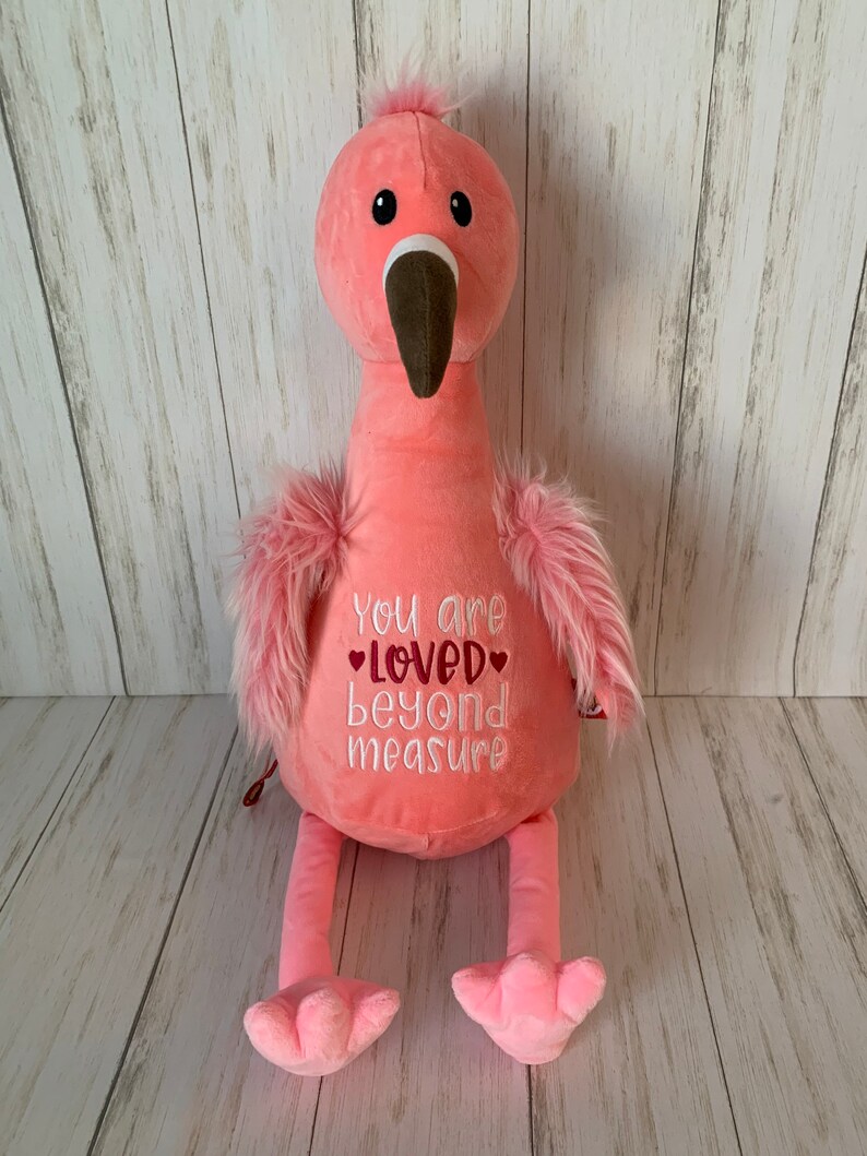May include: A soft pink flamingo toy with a long neck, brown beak, and fluffy pink feathers. The flamingo has the embroidered text "You are loved beyond measure" on its chest. The background is a light wood panel.