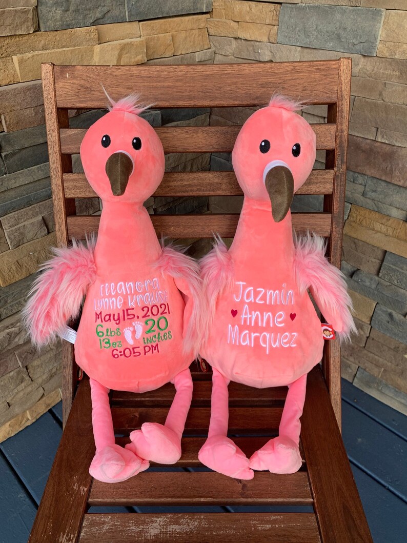 May include: Two pink plush flamingo toys with embroidered details. One flamingo has the name "Eleanora Lynne Krause" and birth details. The other flamingo has the name "Jazmin Anne Marquez". Both are seated on a wooden chair.