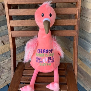 May include: A pink plush flamingo toy with a brown beak and fluffy pink feathers. The flamingo has embroidered details including the name "Eleanor", birth date, weight (410 g), length (49.5 cm), and time of birth.