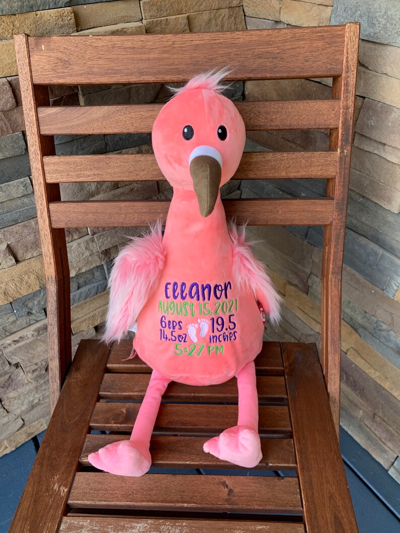 May include: A pink plush flamingo toy with a brown beak and fluffy pink feathers. The flamingo has embroidered details including the name "Eleanor", birth date, weight (410 g), length (49.5 cm), and time of birth.