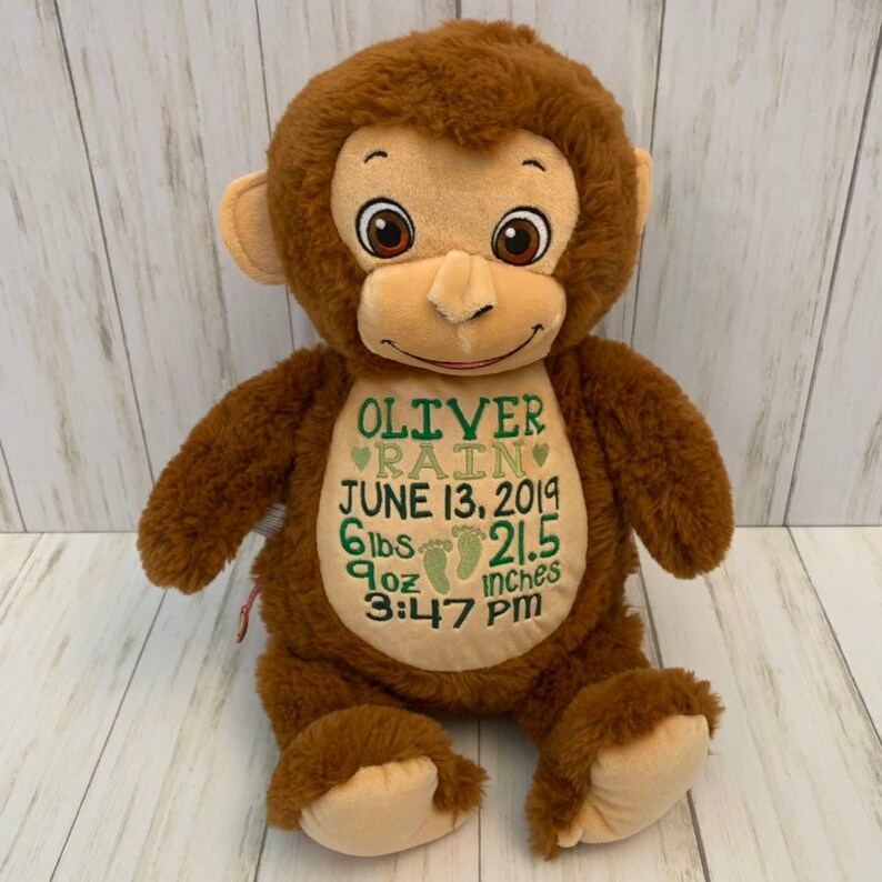 May include: A brown plush monkey with a light brown face, ears, and hands. The monkey has embroidered details and the name "OLIVER RAIN" with birth details: June 13, 2019, 6 lbs, 9 oz, and 21.5 inches.