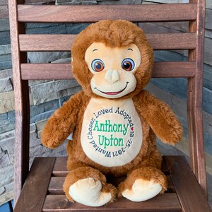 May include: A brown plush monkey doll with a smiling face, sitting on a wooden chair. The doll has a cream-colored face, hands, and feet. The doll has the text "Chosen, Loved, Adopted" around the name "Anthony Upton" and the date "10-25-2021".
