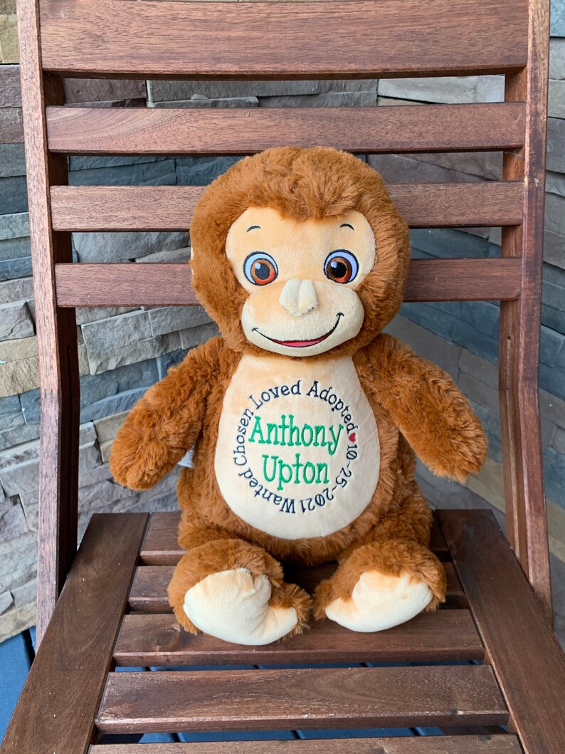 May include: A brown plush monkey doll with a smiling face, sitting on a wooden chair. The doll has a cream-colored face, hands, and feet. The doll has the text "Chosen, Loved, Adopted" around the name "Anthony Upton" and the date "10-25-2021".