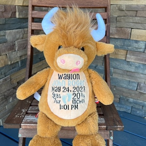 May include: A plush brown Highland cow stuffed animal with light blue horns, sitting on a wooden chair. The cow has embroidered details and a pink tongue sticking out. Text on the cow reads "Waylon John Roger, May 24, 2023, 9lbs 7oz, 20 3/4 inches, 1:01 PM."