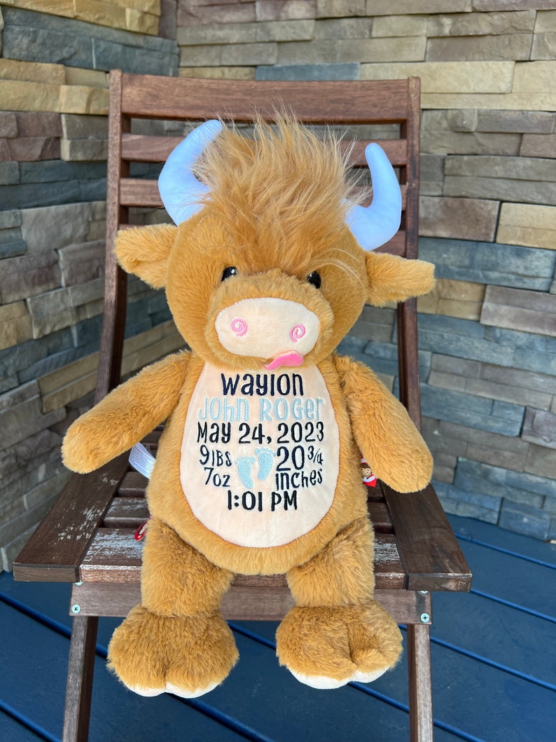 May include: A plush brown Highland cow stuffed animal with light blue horns, sitting on a wooden chair. The cow has embroidered details and a pink tongue sticking out. Text on the cow reads "Waylon John Roger, May 24, 2023, 9lbs 7oz, 20 3/4 inches, 1:01 PM."