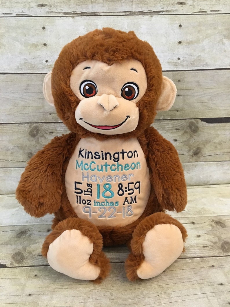 May include: A plush brown monkey with embroidered details. The monkey has a tan face, ears, and feet. Text on the monkey's belly includes a name, date, time, weight, and height: 5 lbs 18 inches 8:59 AM.