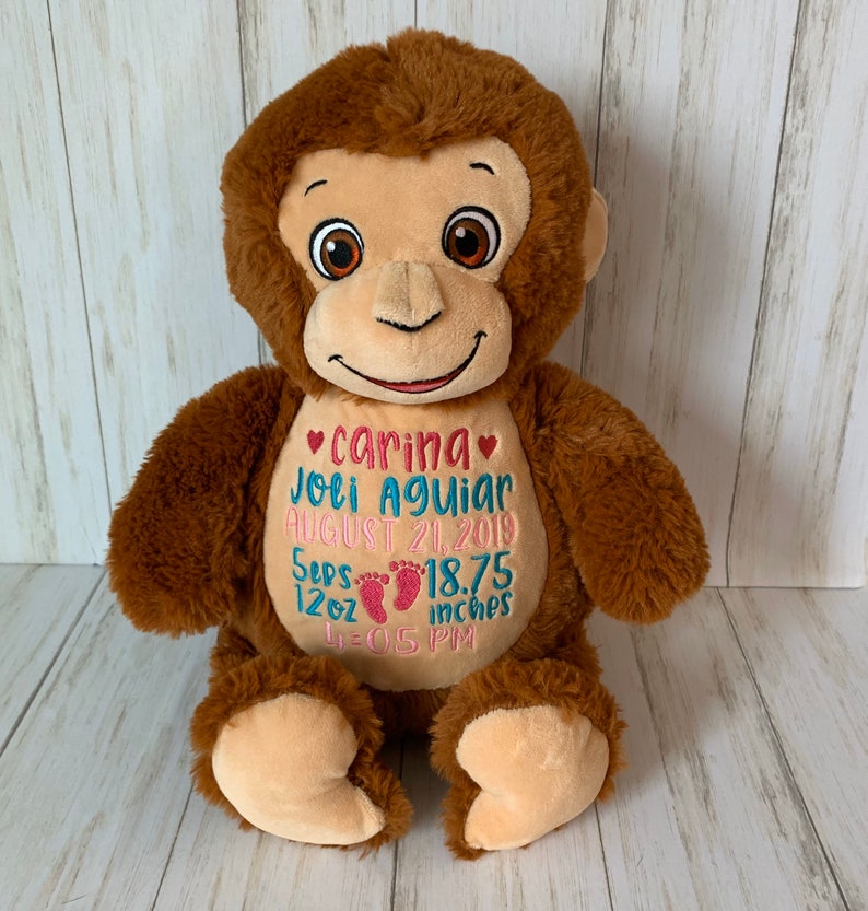 May include: A plush brown monkey with a tan face and belly. The monkey has embroidered details, including the name "Carina" and birth details in pink and blue thread. The monkey is a soft toy.