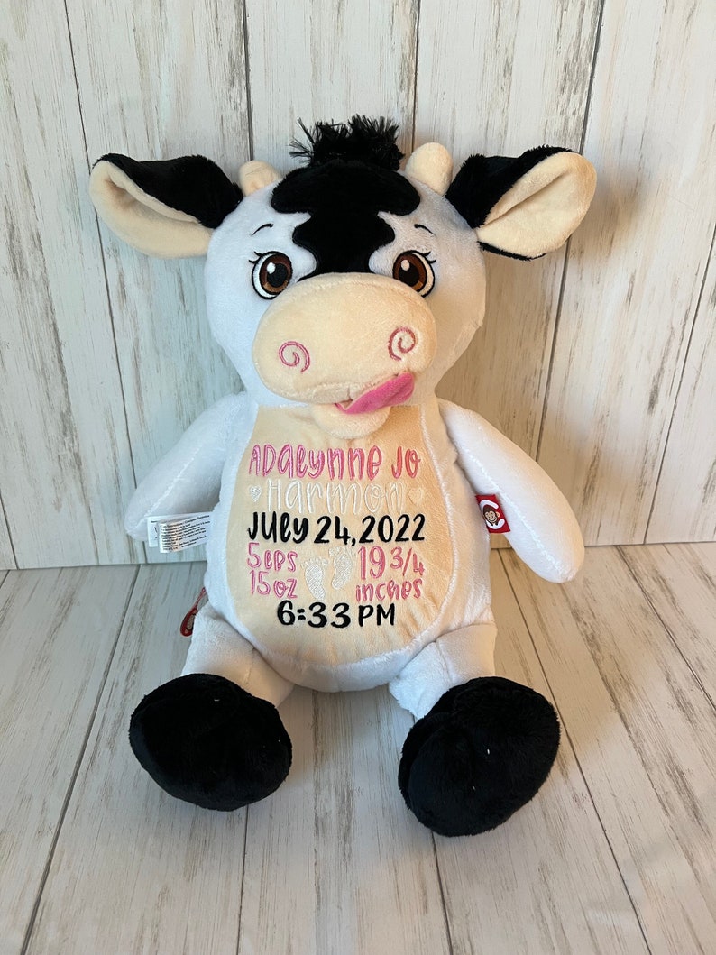 May include: A white plush cow with black patches, ears, and hooves. The cow has embroidered details, including a pink tongue, and text that reads "Adalynne Jo Harmon, July 24, 2022, 5 lbs, 15 oz, 19 3/4 inches, 6:33 PM."