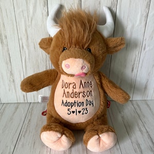 May include: A plush brown Highland cow stuffed animal with white horns and a pink tongue. The cow has embroidered text on its belly that reads "Dora Anne Anderson Adoption Day 5-1-23".