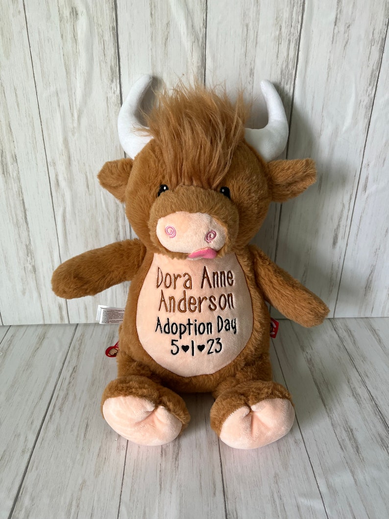 May include: A plush brown Highland cow stuffed animal with white horns and a pink tongue. The cow has embroidered text on its belly that reads "Dora Anne Anderson Adoption Day 5-1-23".