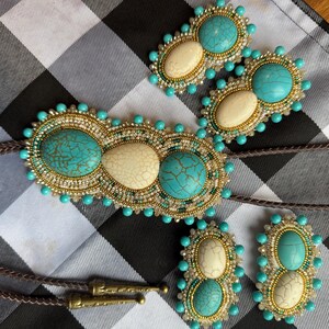 May include: A bolo tie and matching earrings set featuring turquoise and off-white oval stones. The stones are set in a beaded design with gold and turquoise accents. The bolo tie has a brown cord and decorative metal tips.