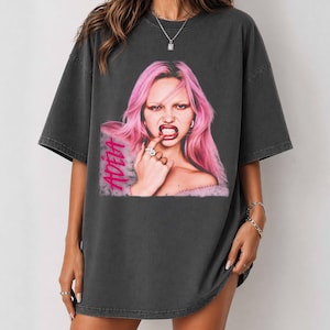 May include: A charcoal grey oversized t-shirt featuring a graphic print of a person with bright pink hair, dark lipstick, and a ring on their finger. The text ADELA is printed in a bold pink font on the left side of the graphic design.