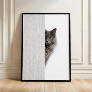 Pode incluir: Impressão artística emoldurada com um gato cinzento de olhos verdes espreitando por trás de um painel branco texturizado. O gato tem pelo fofo, e a obra de arte está emoldurada numa moldura preta. A impressão é exibida num chão de madeira.