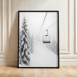 Op de afbeelding: Een zwart ingelijste kunstdruk met een zwart-witfoto van een skiliftstoeltje dat aan kabels boven een besneeuwde berghelling hangt. De scène bevat hoge, met sneeuw bedekte naaldbomen en een mistige, bewolkte winterlucht.
