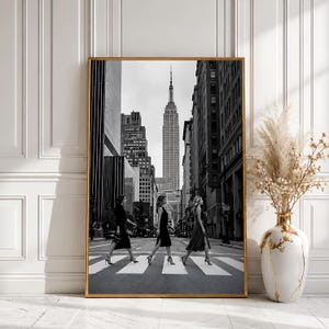 Könnte beinhalten: Ein rechteckiger Wandkunstdruck in einem dünnen goldenen Rahmen mit einer Schwarz-Weiß-Fotografie von drei Personen, die über einen Zebrastreifen gehen, wobei das Empire State Building im Hintergrund zwischen hohen Wolkenkratzern zu sehen ist.
