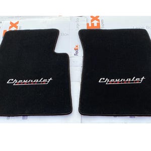 May include: Two black car floor mats with the Chevrolet Impala 1960 logo embroidered in white and red. The mats are rectangular with a custom fit and a textured surface.