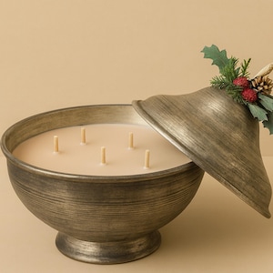 May include: A large, antique-style candle in a bronze-colored bowl with a matching lid. The candle has six wicks and is decorated with a sprig of holly, berries, and a pinecone on the lid. The candle is a decorative home accent.