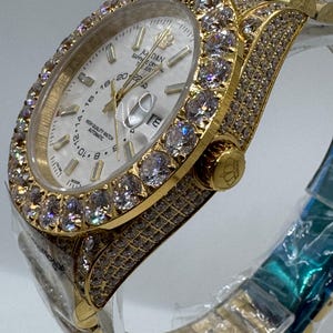 Puede incluir: Un reloj de pulsera dorado con esfera blanca y bisel y correa incrustados con piedras preciosas transparentes. La esfera del reloj muestra la hora y la fecha. El reloj est&aacute; etiquetado como "JORDAN" y "HIGH QUALITY WATCH AUTOMATIC".