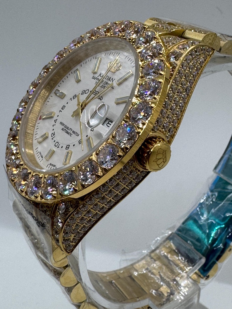 Puede incluir: Un reloj de pulsera dorado con esfera blanca y bisel y correa incrustados con piedras preciosas transparentes. La esfera del reloj muestra la hora y la fecha. El reloj est&aacute; etiquetado como "JORDAN" y "HIGH QUALITY WATCH AUTOMATIC".